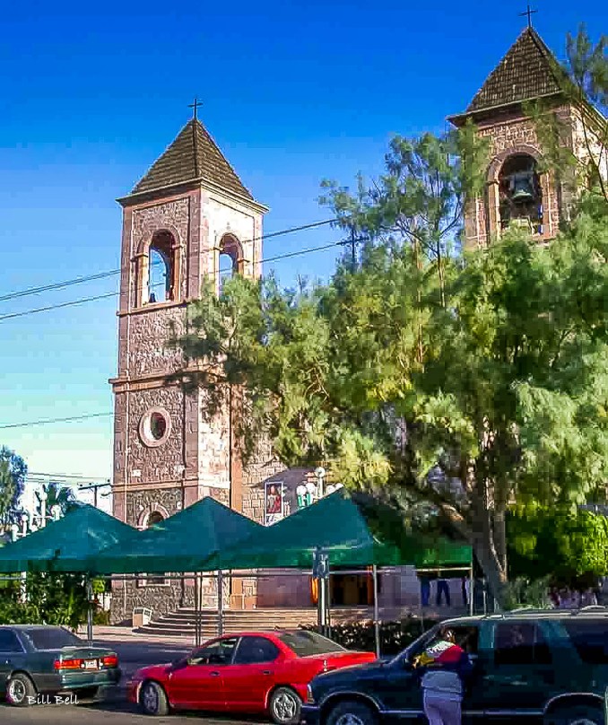 The twin bell towers of the Catedral de Nuestra Señora de La Paz, standing tall in the heart of La Paz, Baja California Sur, framed by lush greenery and a bright blue sky.