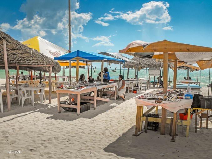 A vibrant market scene on the sandy shores of Celestún, where locals sell crafts and fresh seafood under colorful canopies, with the turquoise waters of the Gulf of Mexico in the background.