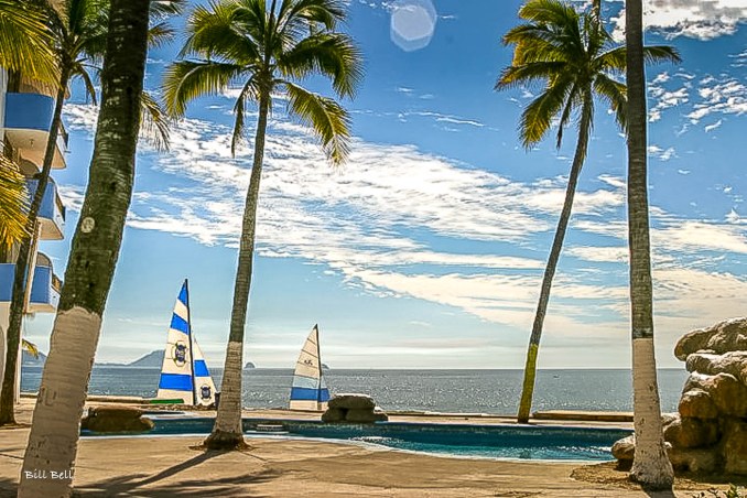 Colorful sailboats dot the waterfront at a resort in Mazatlán, with palm trees framing the idyllic scene of sun, sand, and sea.