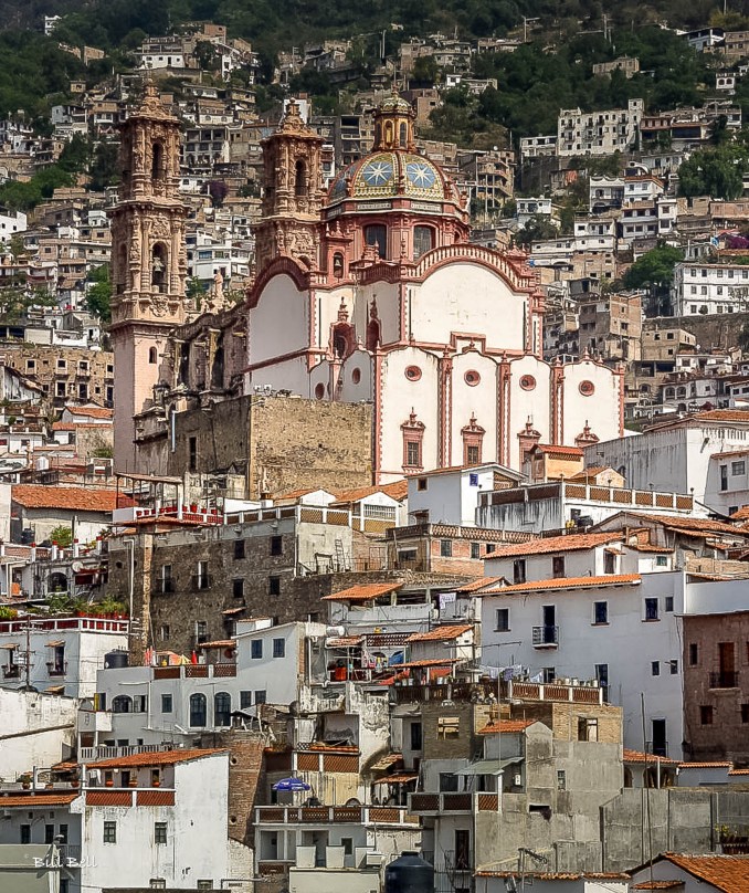 The Santa Prisca Church, seen from a distance, framed by the lush hillside and white buildings of Taxco. This iconic view captures the beauty and elevation of the town, perched in the hills of Guerrero.