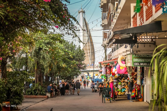 A vibrant street in Puerto Vallarta leads to the famous Los Muertos Pier, bustling with shops, restaurants, and local artisans. The lush greenery and colorful storefronts make for a perfect stroll through this picturesque city