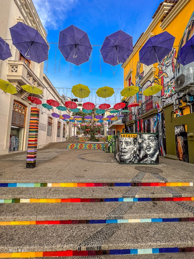 A colorful, artsy alleyway in Puerto Vallarta, adorned with hanging umbrellas and street murals. This lively scene is a testament to the city’s thriving artistic community and its vibrant street art culture.