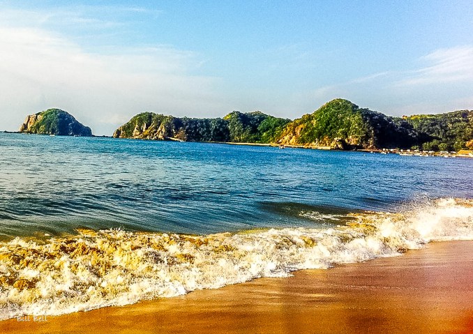 The golden sands of Barra de Navidad meet the clear blue waters of Bahía de Navidad, with lush green hills in the distance. This picturesque view is a perfect representation of the natural beauty that attracts visitors to the area.