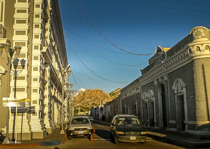 A picturesque street in Hermosillo, lined with historic buildings, leads the eye towards the scenic hills that frame the city, highlighting the city's unique charm.