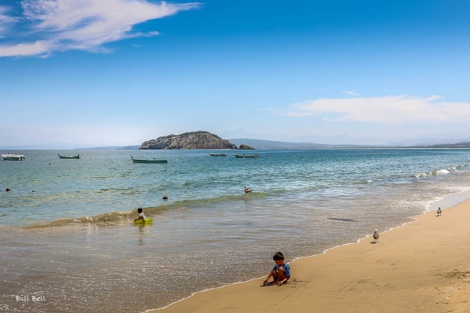 Children enjoying the warm waters of Los Ayala beach, with the scenic backdrop of small fishing boats and distant islands.