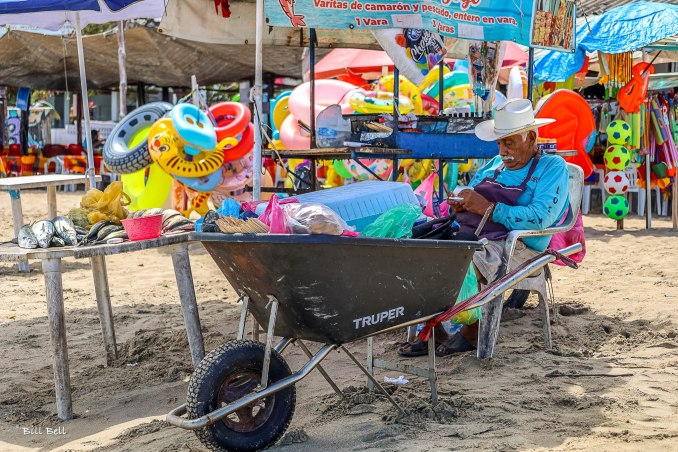 A local vendor resting under the shade at Los Ayala beach, selling fresh fish and a variety of beach toys to tourists.