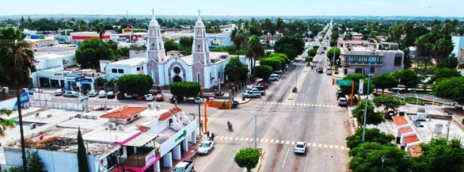 Aerial view of the central area of Huatabampo, Sonora, showcasing its iconic twin-towered church and the main street lined with lush greenery and local businesses