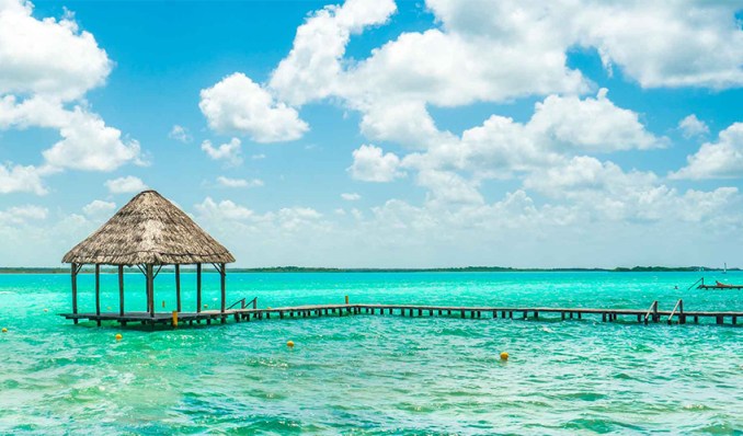 A picturesque view of a thatched-roof pier extending into the turquoise waters of Chetumal Bay. The clear blue sky and serene water create a perfect setting for relaxation and enjoyment in Chetumal, Quintana Roo.