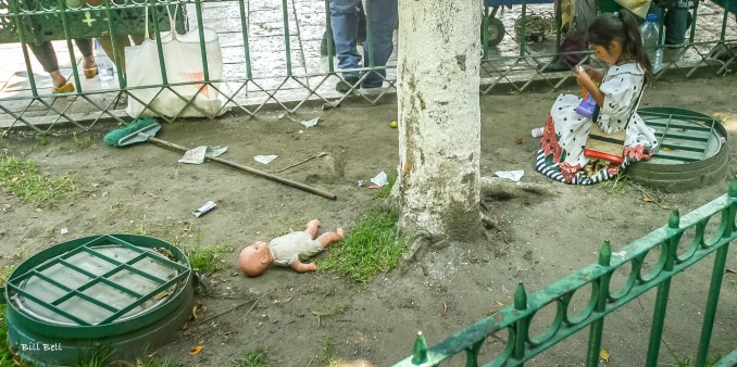 A quiet moment in a busy park as a young girl focuses on her toy, while nearby, a forgotten doll lies abandoned on the ground, telling its own silent story.