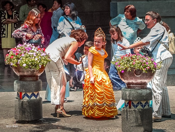 "A young girl dressed in a stunning yellow gown enjoys her moment of royalty in Puebla, surrounded by family and friends in a picturesque plaza.