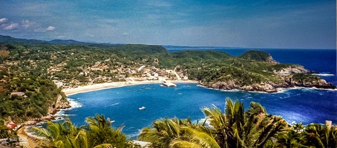 A stunning panoramic view of Puerto Ángel’s serene bay, where turquoise waters meet the lush, green coastline. Fishing boats dot the water, and the village’s charming, rustic houses blend into the hillside, offering a perfect escape into nature along the Oaxaca coast.